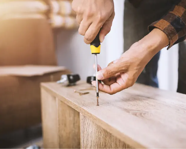 Hände drehen Schraube in Holzplatte ein, daneben Werkzeug und Bauteile auf hellem Untergrund in einer Montagearbeit.